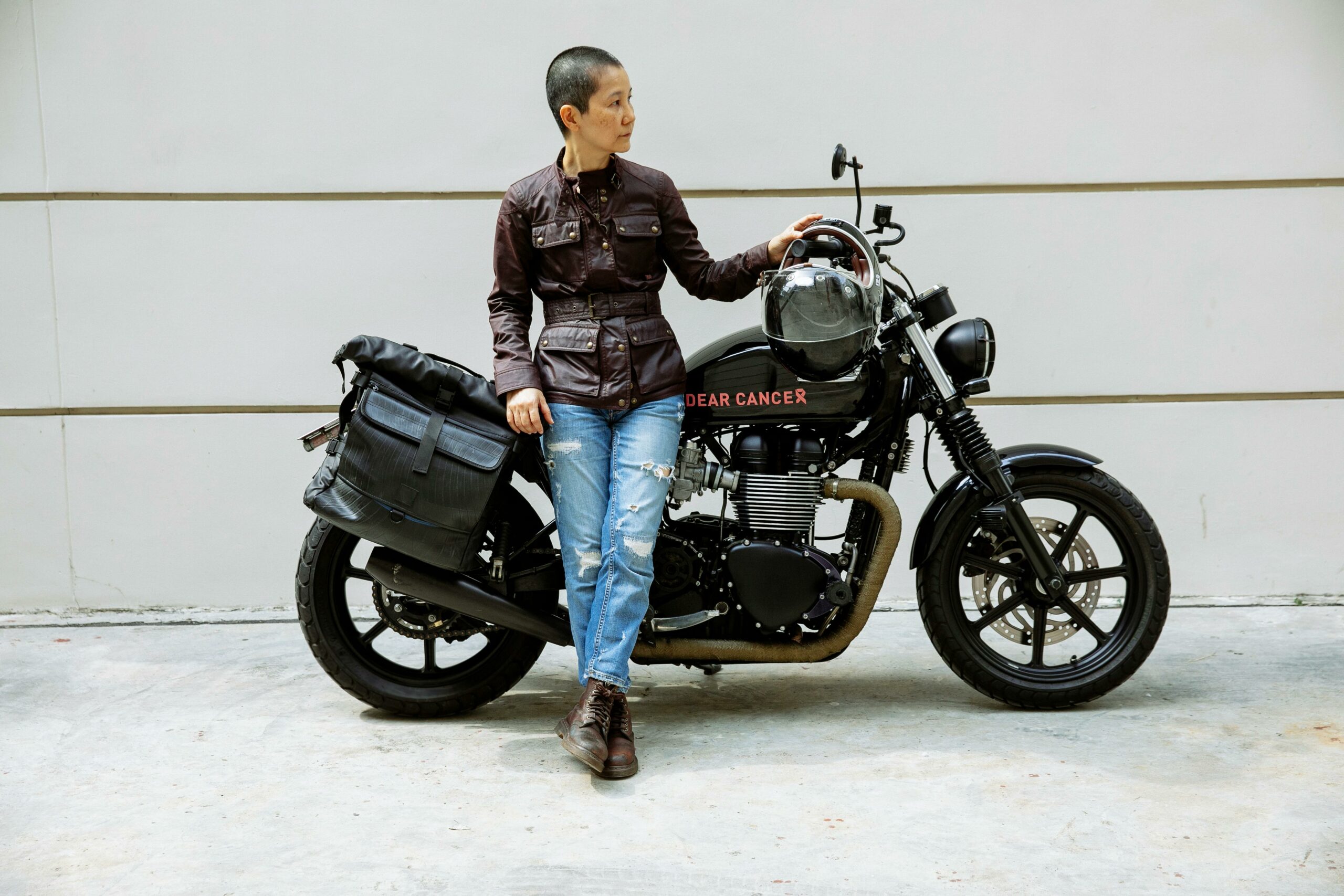 Serious middle age ethnic female biker in jacket and jeans looking away while leaning on black motorbike on asphalt road near wall in daylight in city street